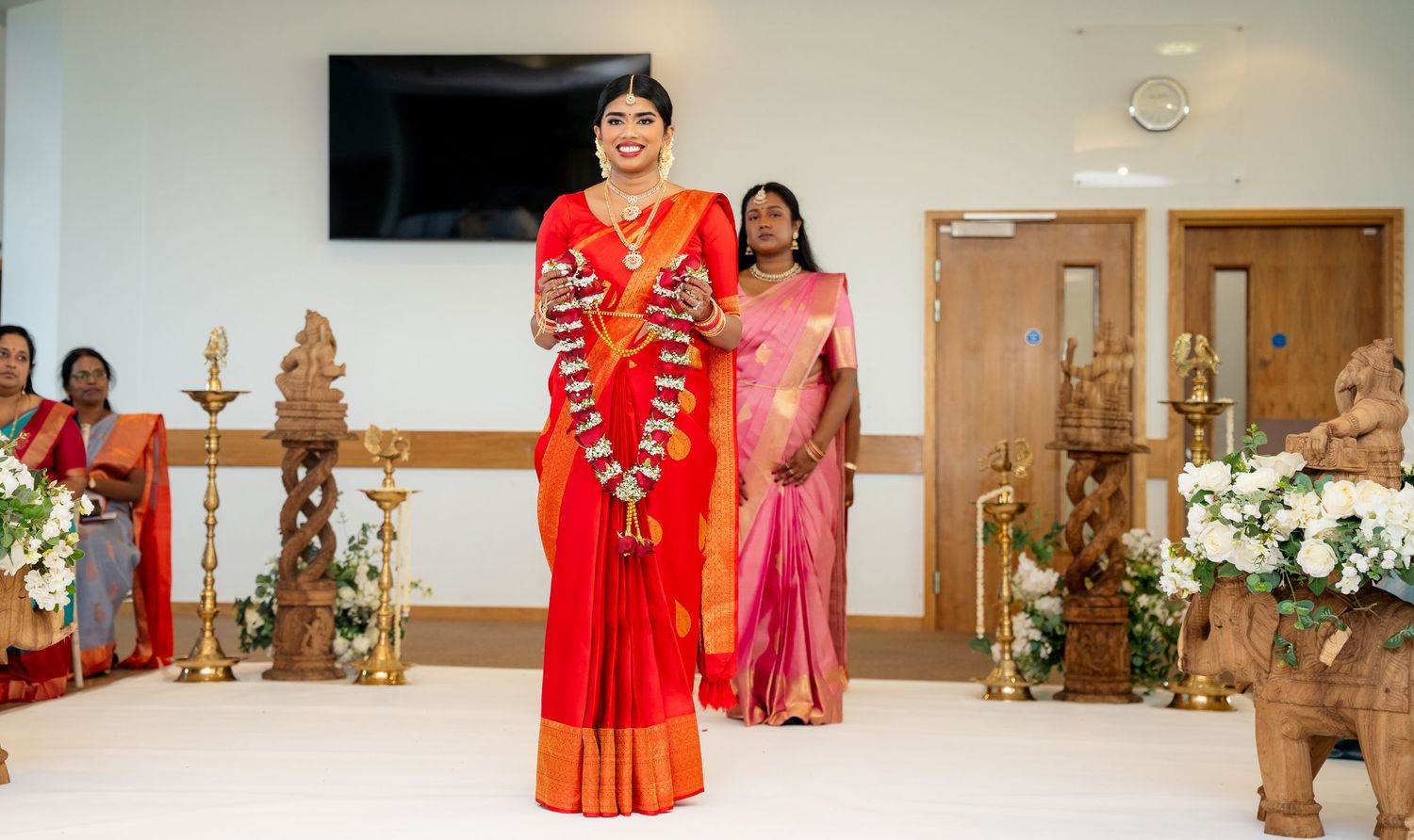 Woman in red saree walks at traditional South Indian wedding ceremony with decorative lamps and floral arrangements.
