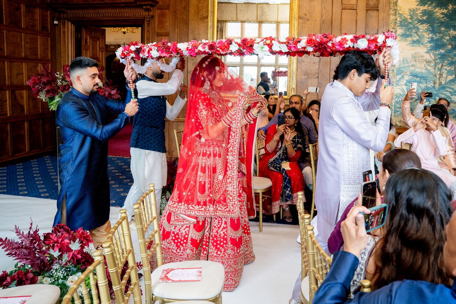 Indian bride in red lehenga entering wedding ceremony under floral mandap with family members.