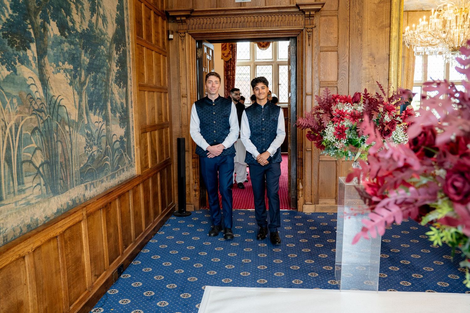 Two men in matching navy vests stand in an elegant wood-paneled room with tapestry and red floral arrangement.