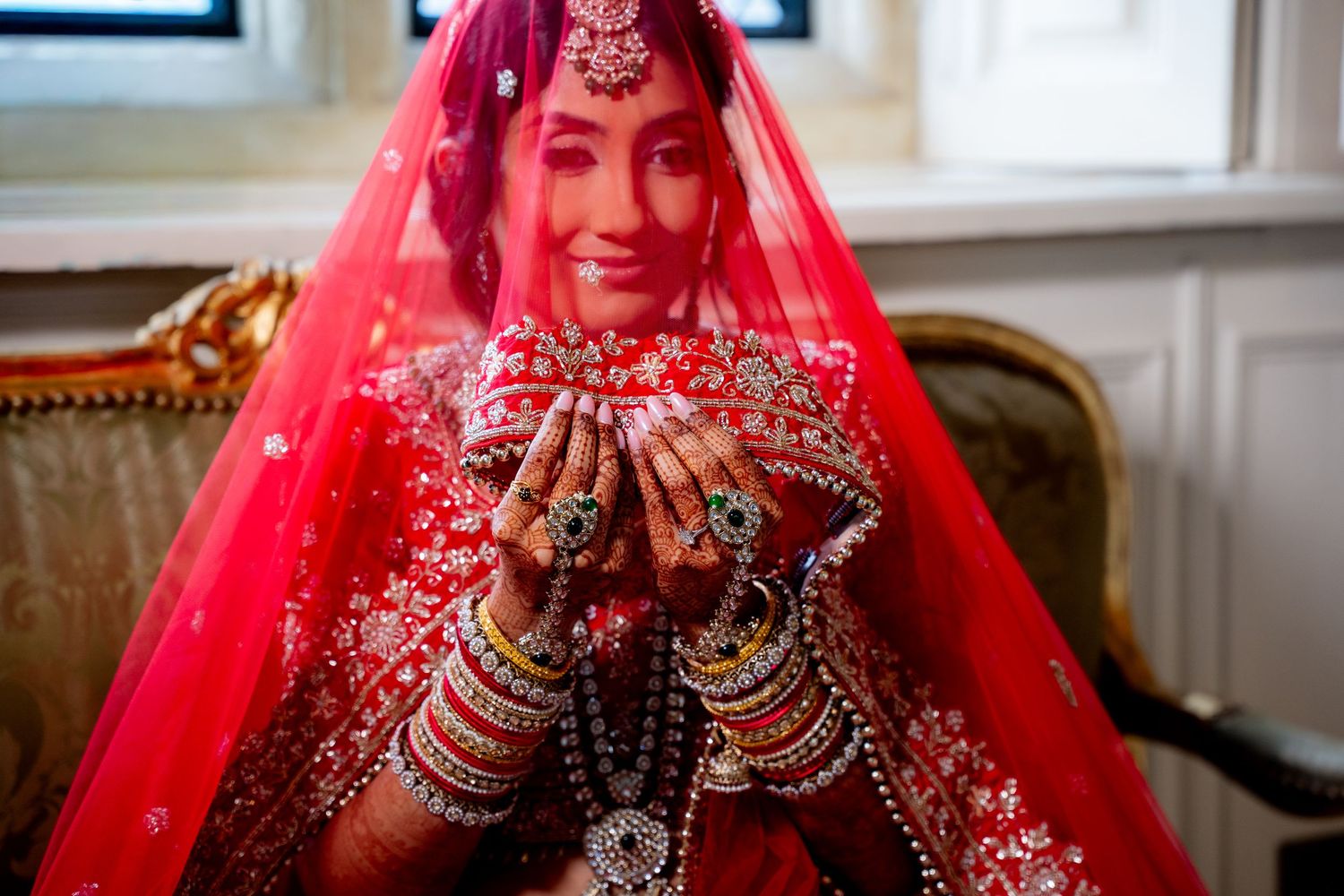 Indian bride in red veil showcasing mehndi hands and colorful bangles with a smile.
