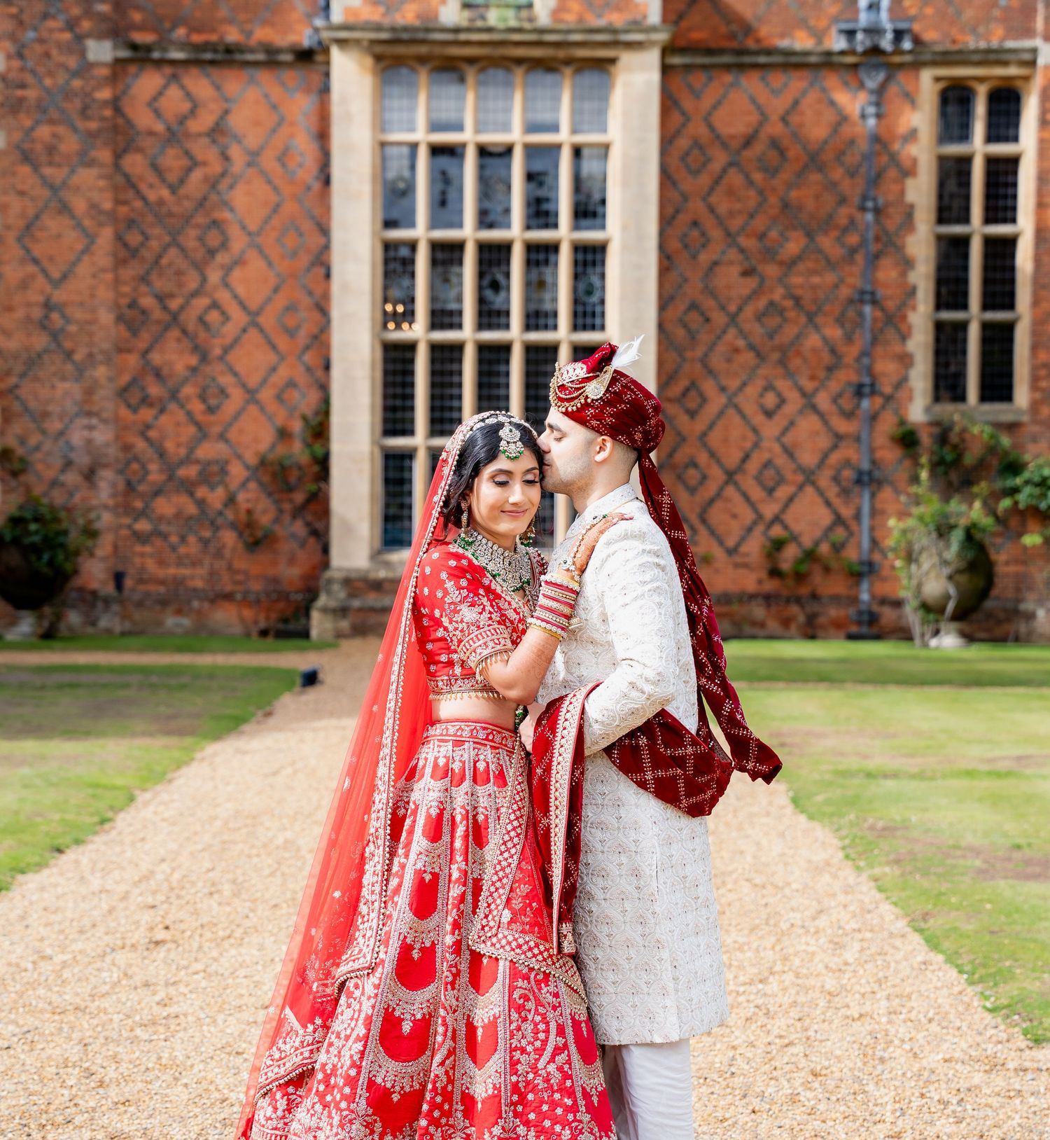 Indian bride and groom in red and white traditional wedding attire pose romantically outside a historic brick manor.