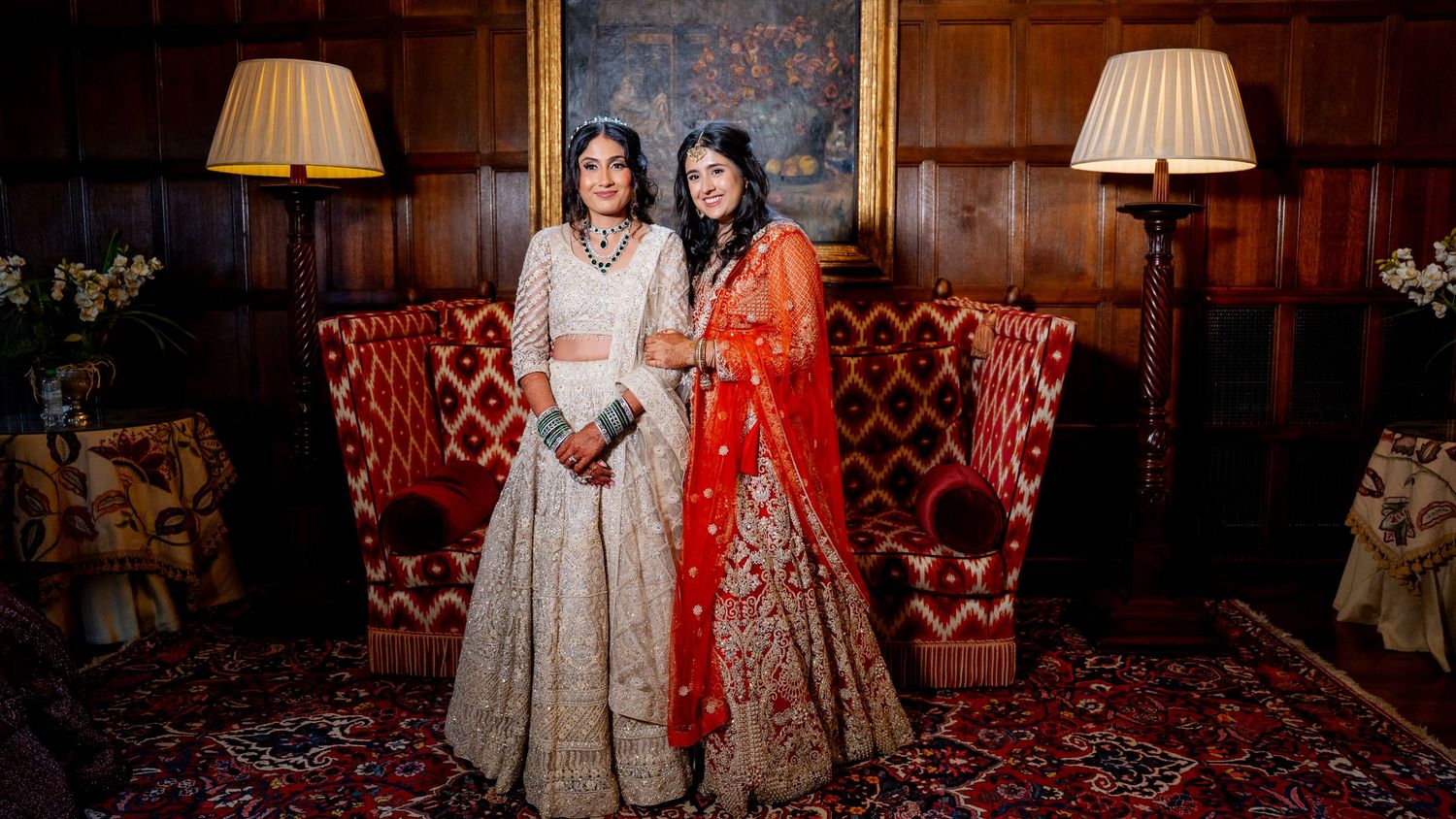 Two women in stunning Indian bridal lehengas pose together in a grand wood-paneled room.