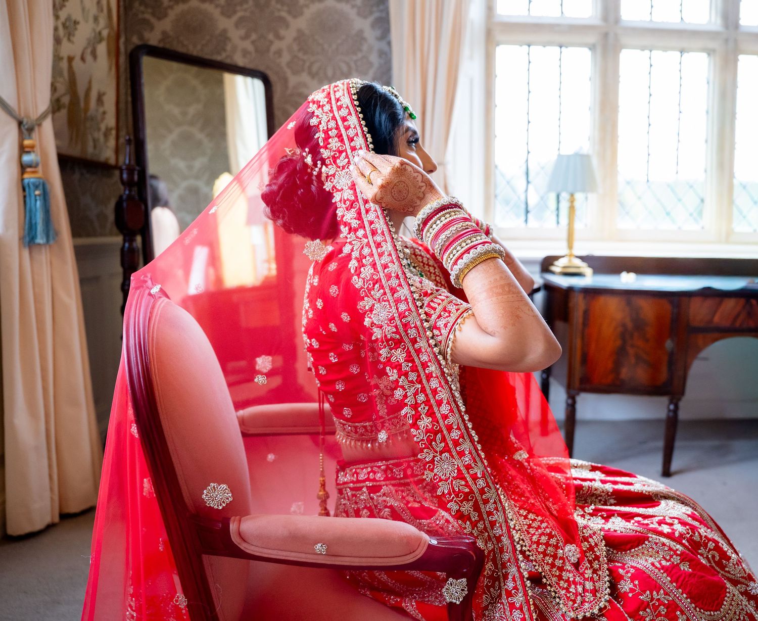 Indian bride in red bridal lehenga and dupatta sitting in chair adjusting jewelry during wedding preparations.