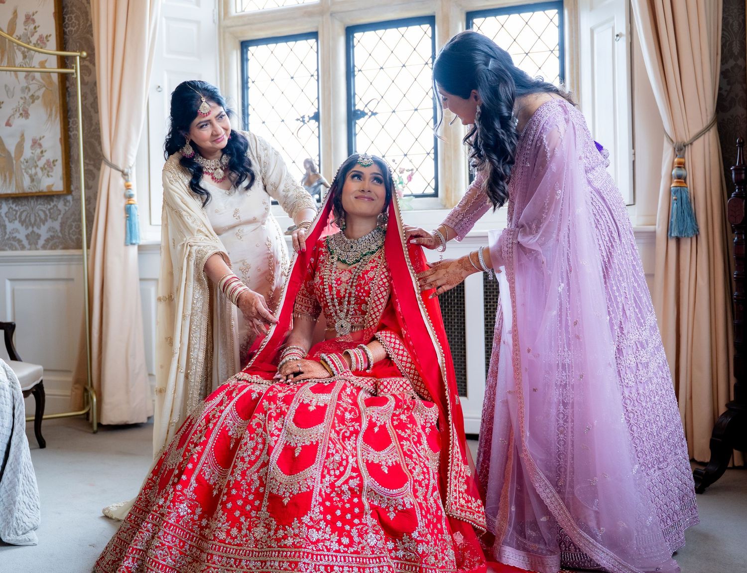 Two women helping Indian bride in red lehenga with final preparations before ceremony.