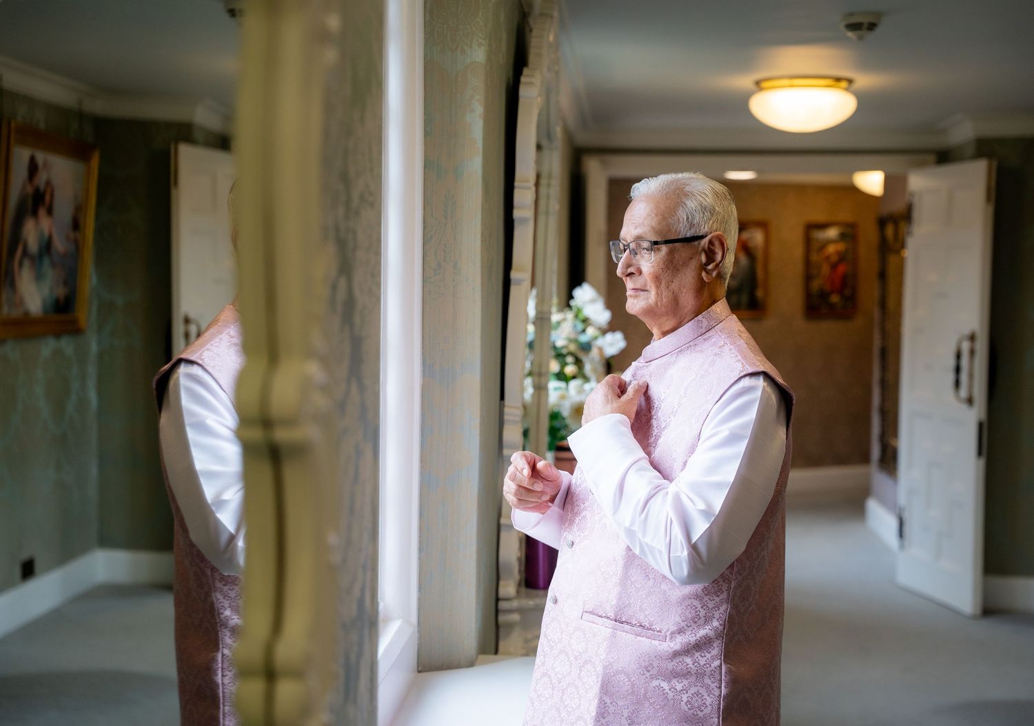 Elderly man in pink kurta adjusting collar while looking in mirror in elegant hallway.