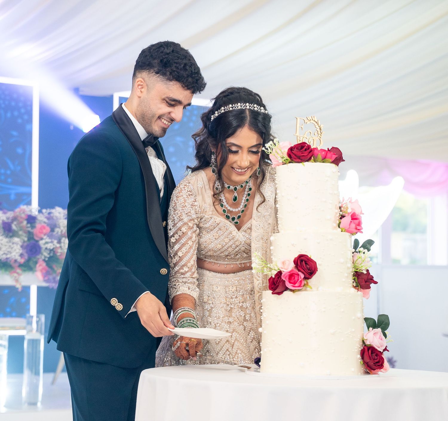 Joyful couple in South Asian wedding attire cutting tall white rose-adorned wedding cake at reception.