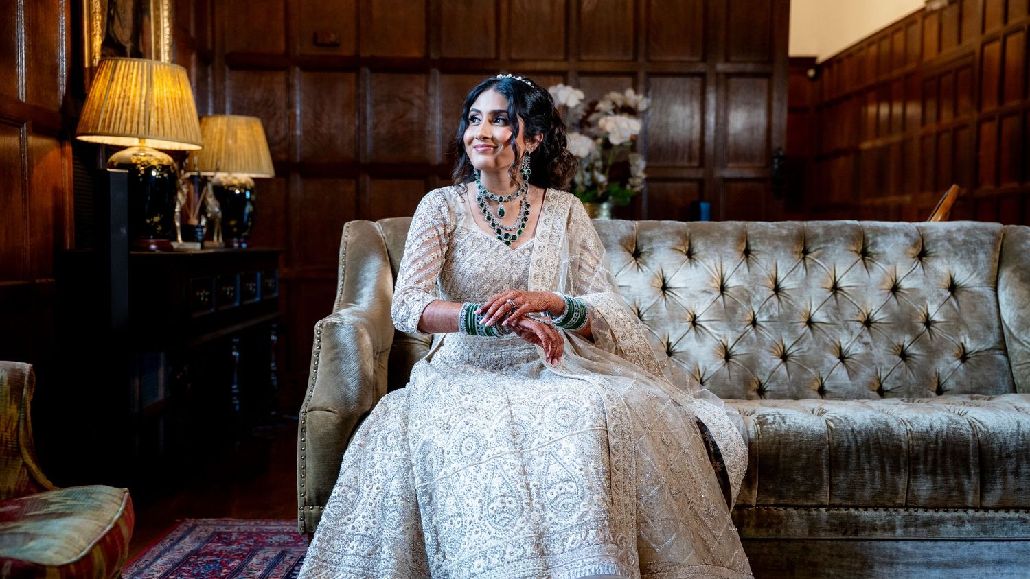 Elegant bride in embroidered lehenga seated on vintage sofa in wood-paneled room with warm lamp lighting.