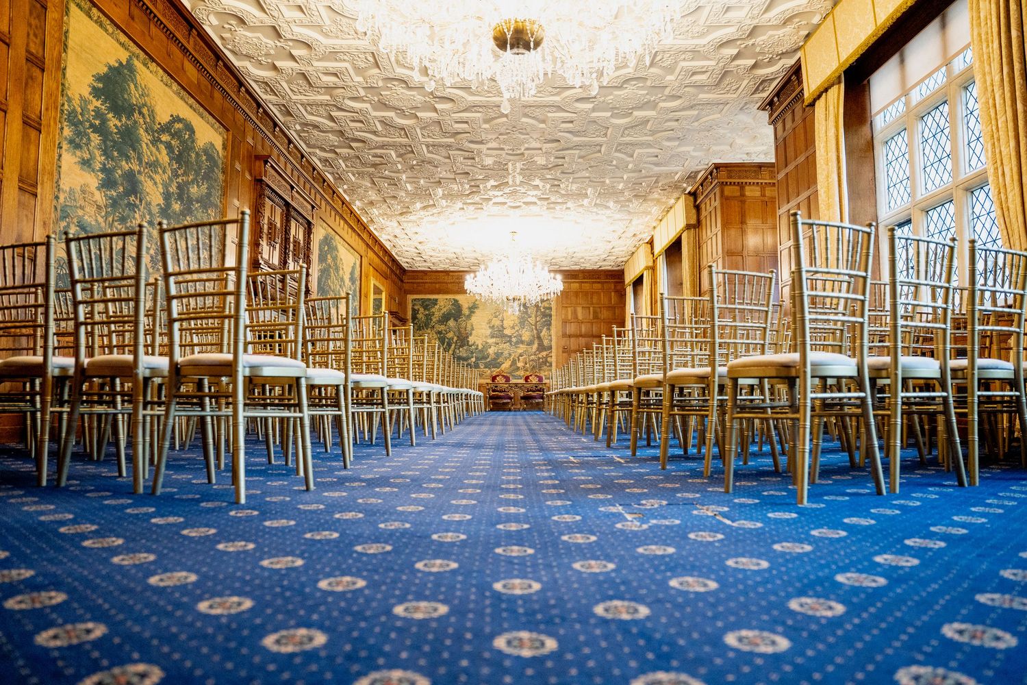 Elegant ballroom with gold Chiavari chairs on blue patterned carpet, ornate ceiling, chandelier, and tapestry wall panels.
