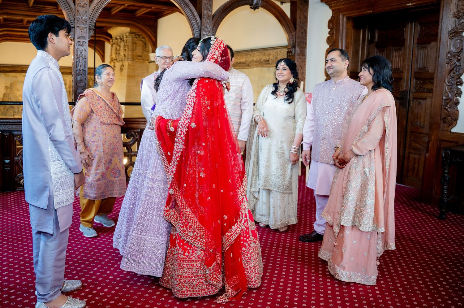 Indian bride in red lehenga embracing family members in ornate wooden hall during wedding ceremony.