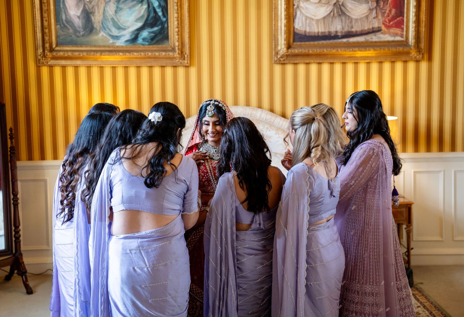 Indian bride surrounded by bridesmaids in lavender sarees in ornate room.