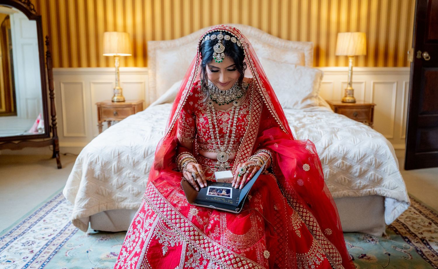Indian bride in red lehenga reading holy book on bed in elegant hotel room before wedding.