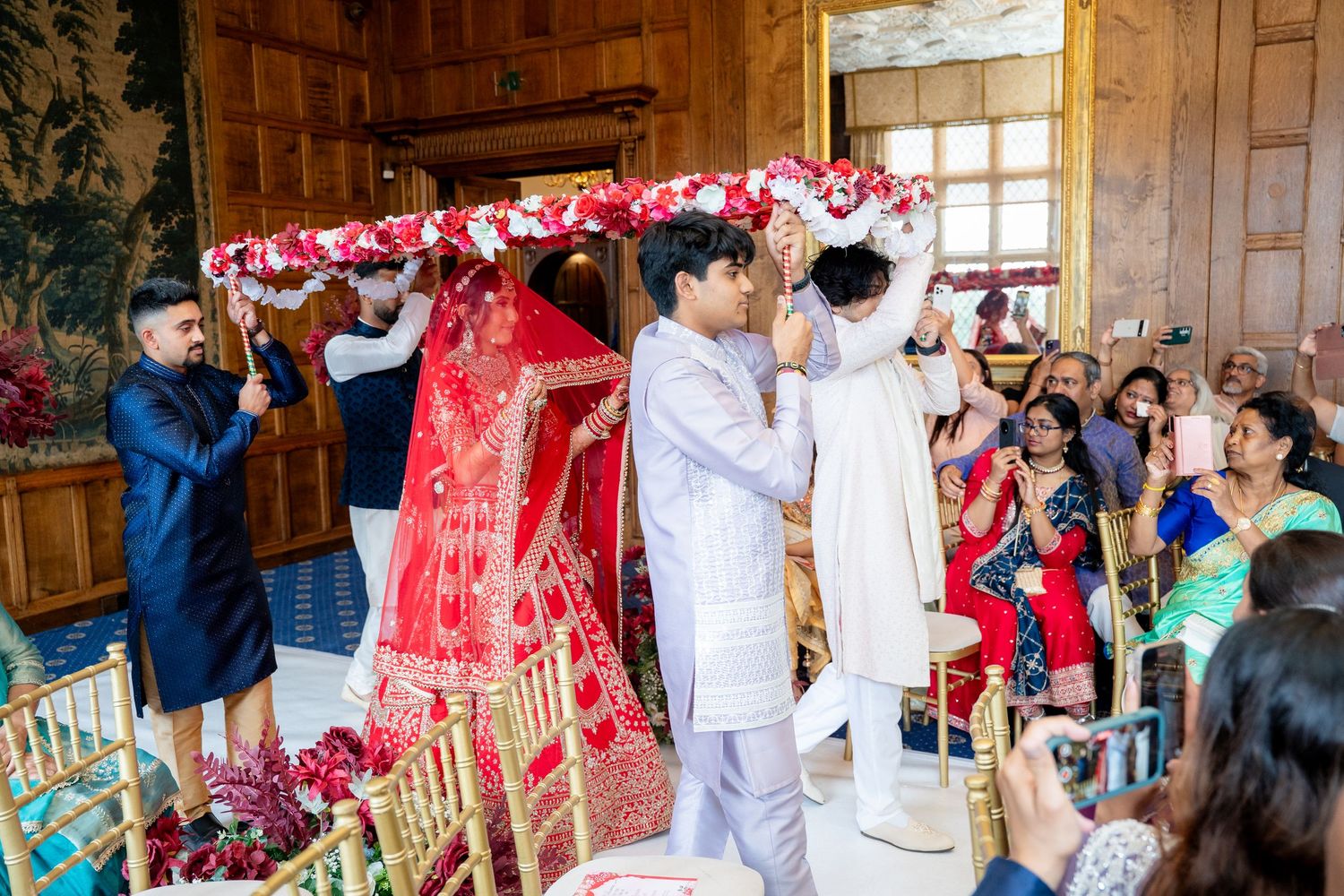 Indian bride in red lehenga enters ceremony under floral canopy held by groomsmen.