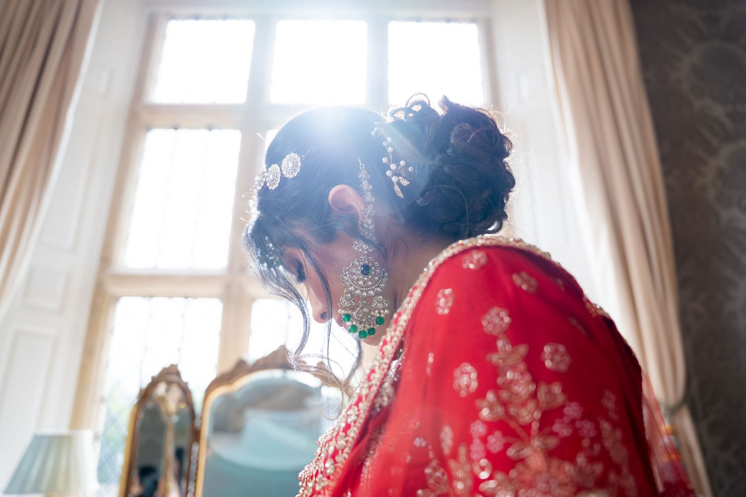 Indian bride in red lehenga with ornate hair accessories and emerald earrings in sunlit room.
