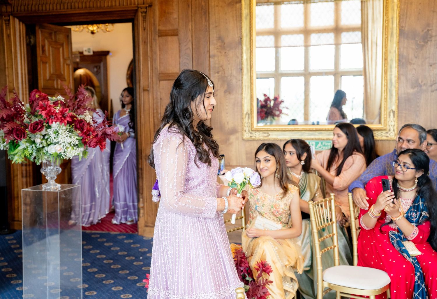 A woman in a pink lehenga walks down the aisle at an Indian wedding ceremony in an elegant wood-paneled venue.