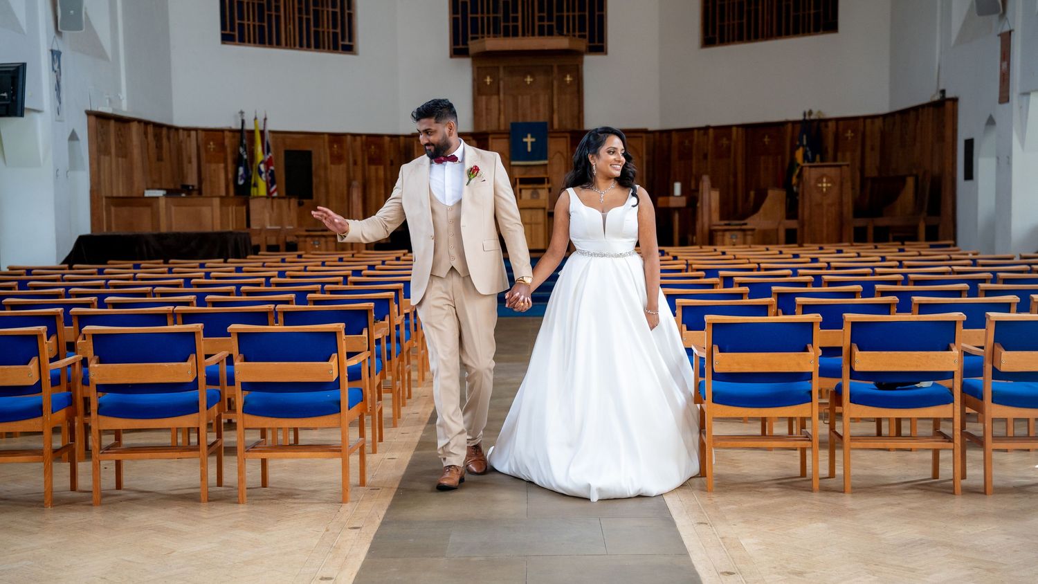 Bride and groom walk hand in hand down the aisle of a church with blue pews after their wedding ceremony.