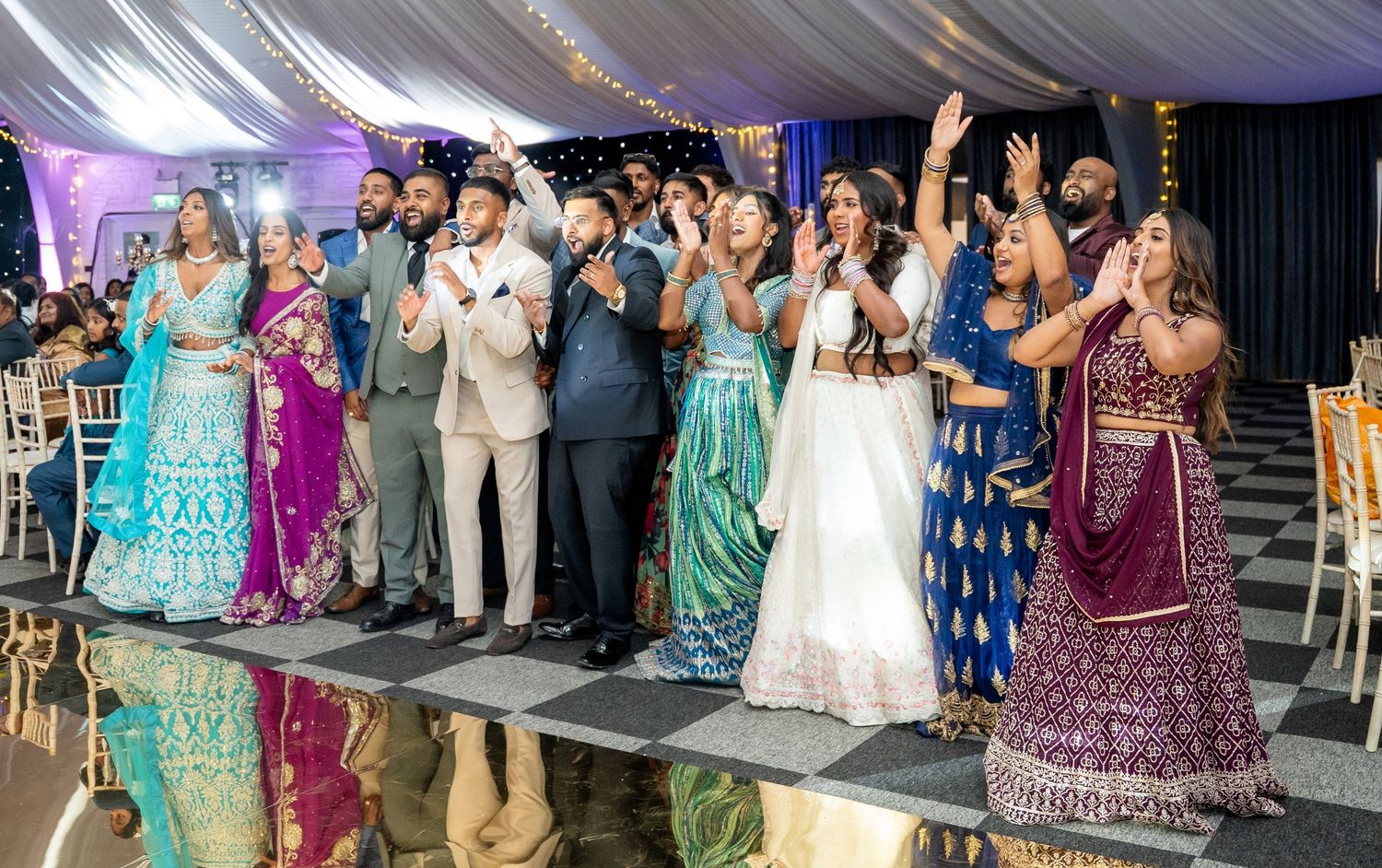 Wedding reception guests celebrating on dance floor in colorful traditional Indian attire and formal wear under draped tent.
