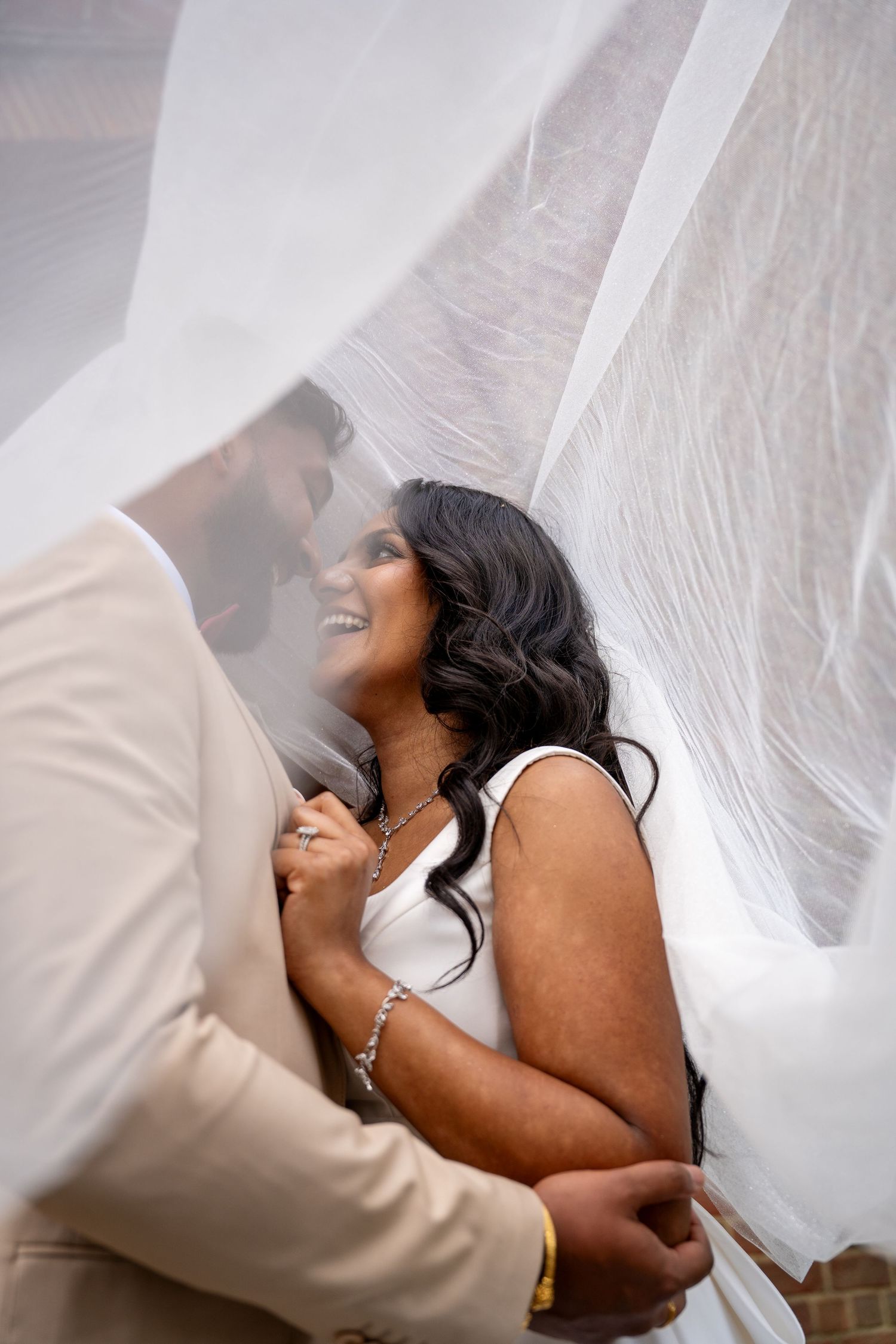 Bride and groom share a romantic moment under a flowing white veil, bride smiling joyfully in a white dress.