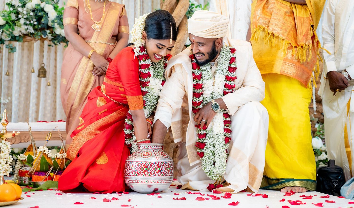 Indian couple in traditional attire performing a Hindu wedding ritual with flower petals and a ceremonial pot.