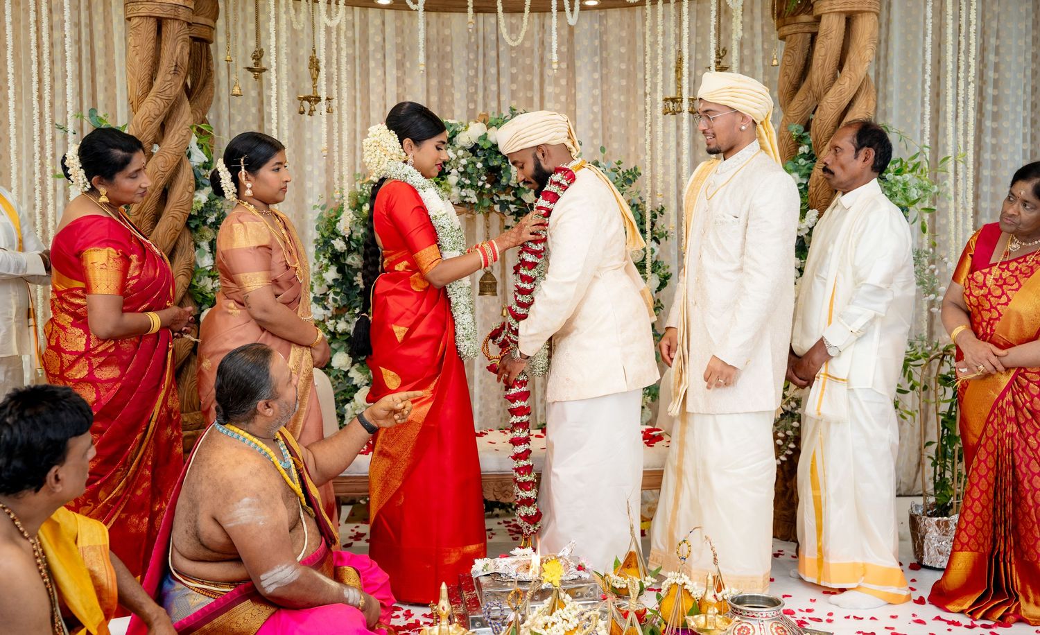 Couple exchanges flower garlands at a colorful South Indian wedding ceremony surrounded by family members.
