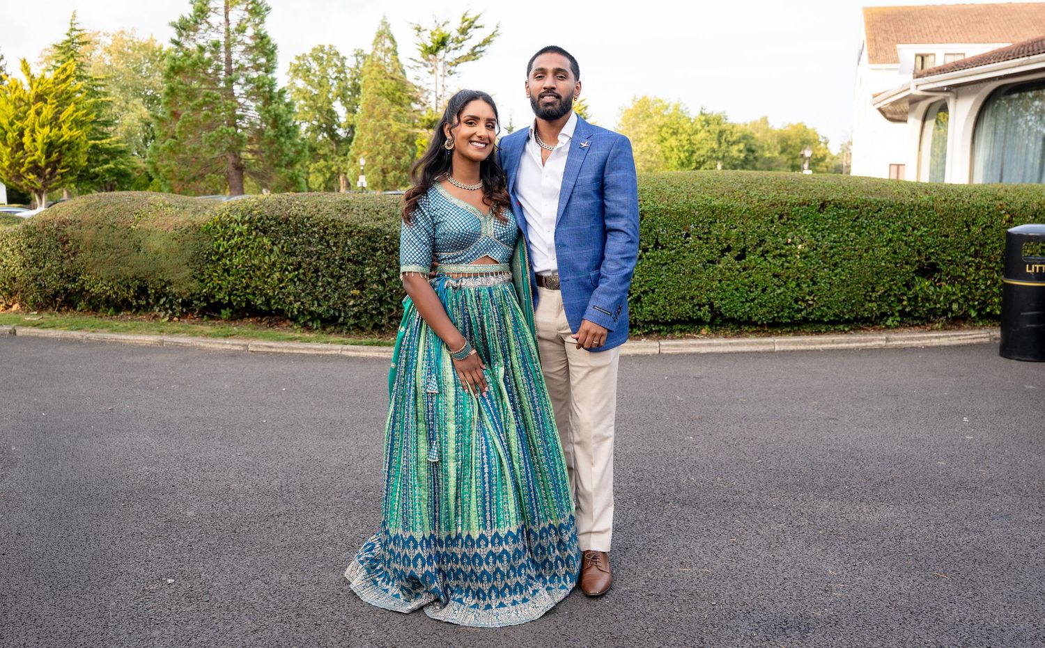 A couple poses together outdoors, the woman in a teal lehenga and the man in a blue blazer.
