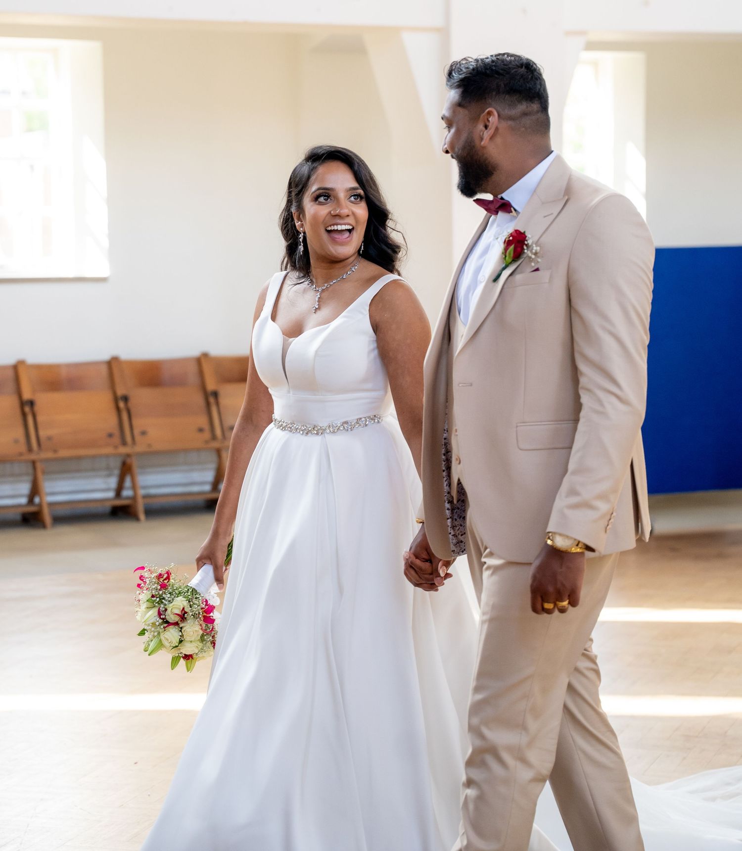 Smiling bride in white gown holds bouquet while walking hand-in-hand with groom in tan suit inside a chapel.