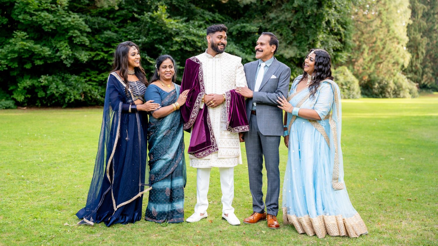Indian wedding family group of five smiling outdoors, wearing traditional attire in blue, white, and purple on green lawn.
