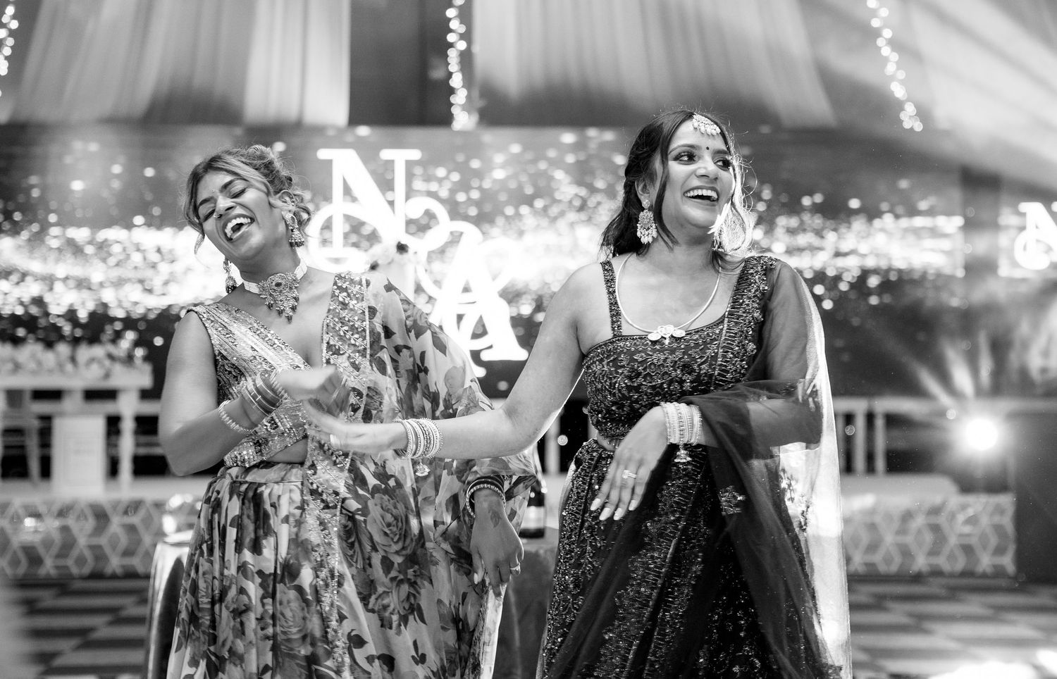 Two women in Indian attire laughing joyfully at a wedding reception with monogram backdrop.