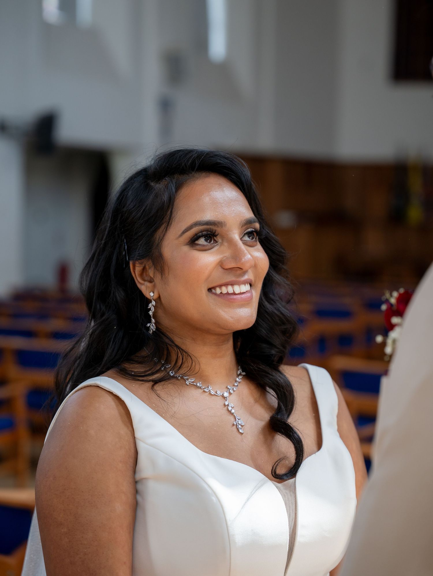 A smiling bride in a white dress wearing a diamond necklace and earrings stands in a church during her wedding ceremony.