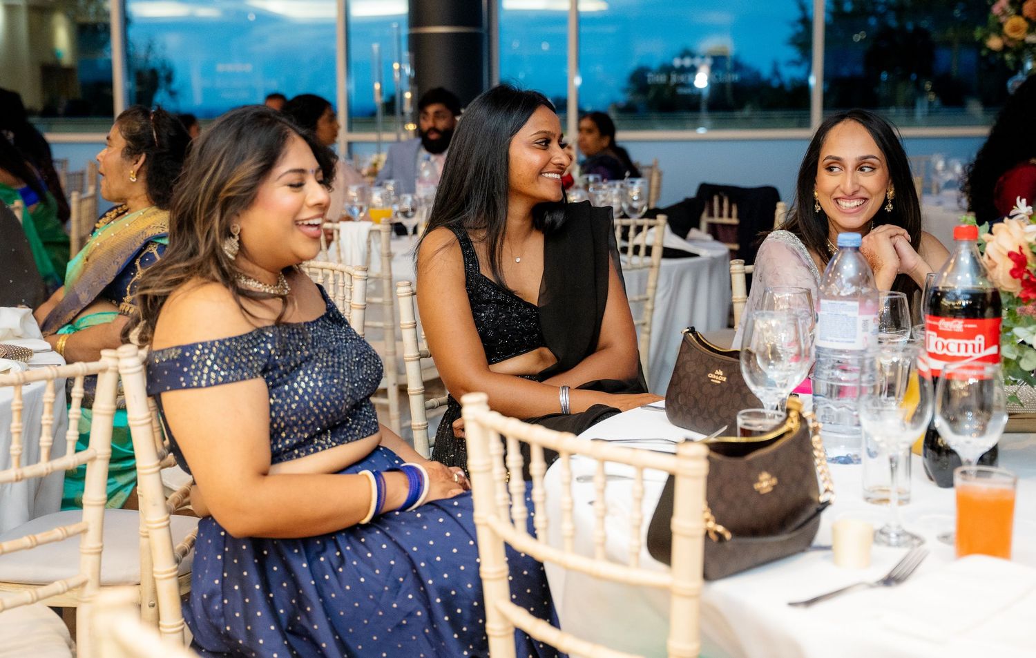 Three women in festive attire laughing together at a decorated banquet dinner table.