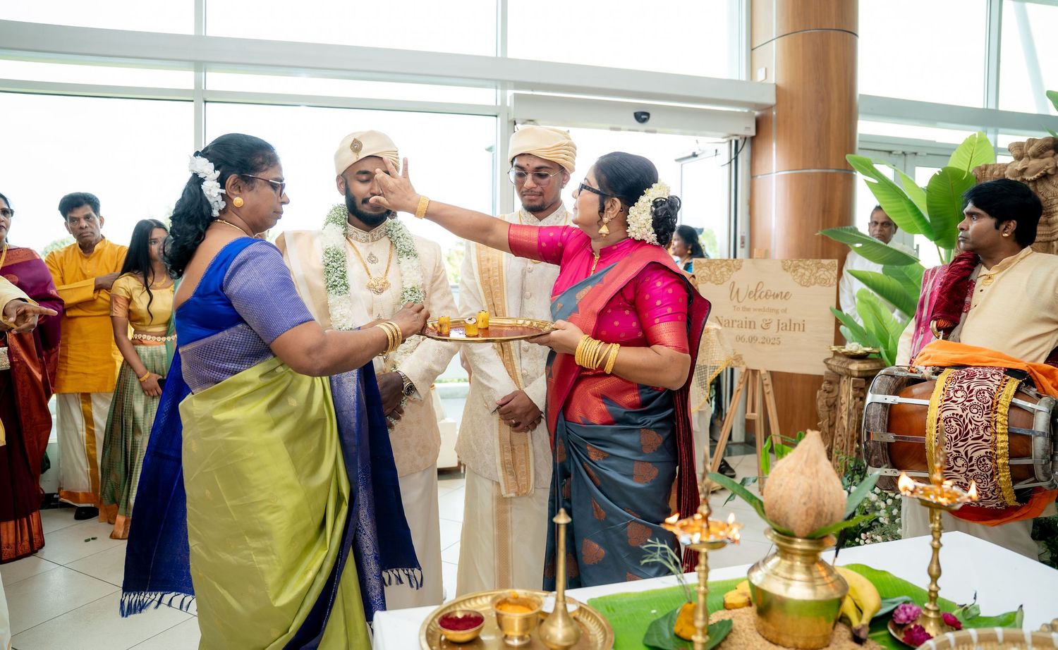 Traditional South Indian wedding ceremony with groom in white turban, guests in colorful sarees, oil lamps, and drum musician.