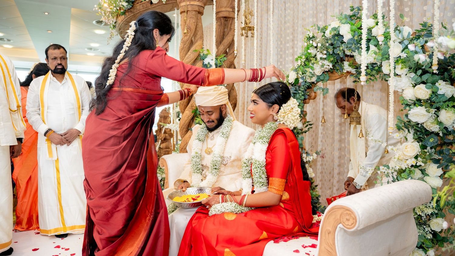 Woman performing traditional blessing ceremony for Hindu bride and groom at decorated Indian wedding mandap.