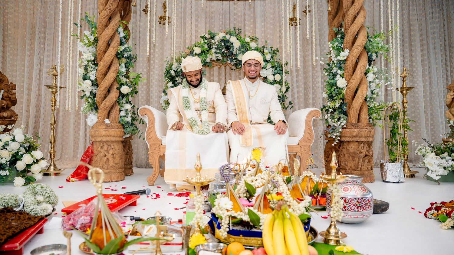 Two grooms in white traditional attire pose on ornate wedding stage surrounded by ritual offerings and fruit.