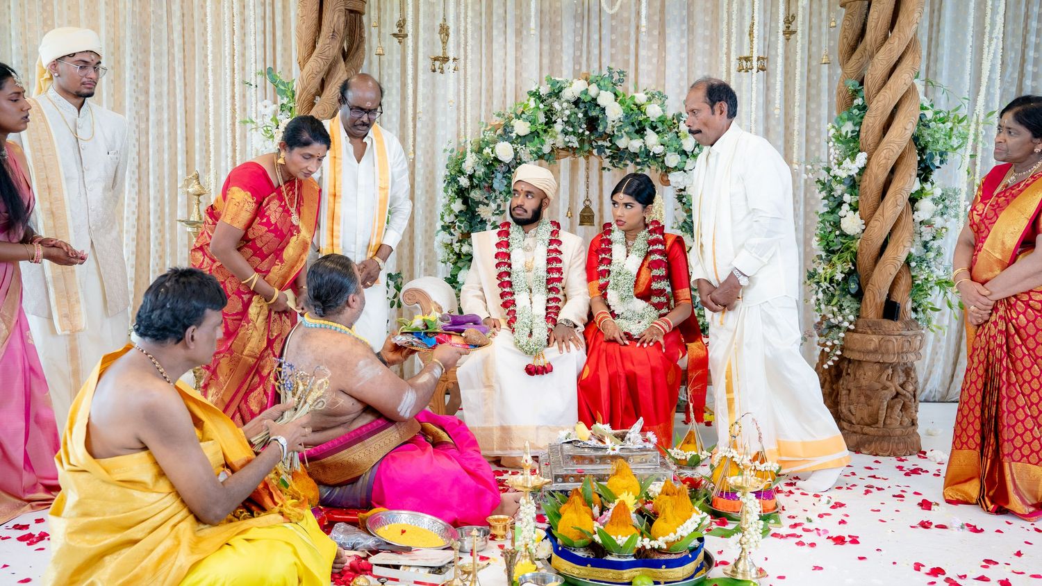 South Indian wedding ceremony with priest, family, bride and groom in traditional attire at decorated mandap altar.