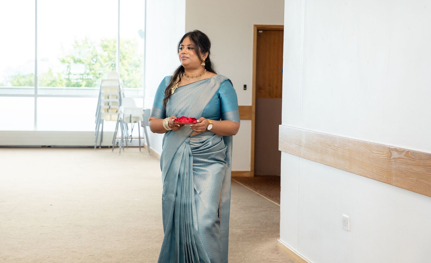 Woman in shimmering teal silk saree carrying decorative red plate at an Indian wedding venue.