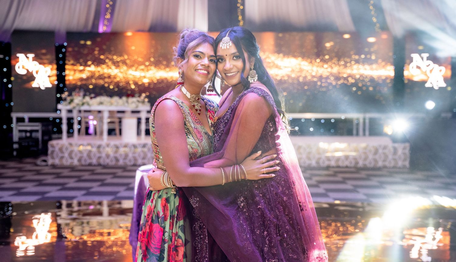 Two women in vibrant Indian lehenga outfits sharing a warm hug on a sparkling wedding dance floor.