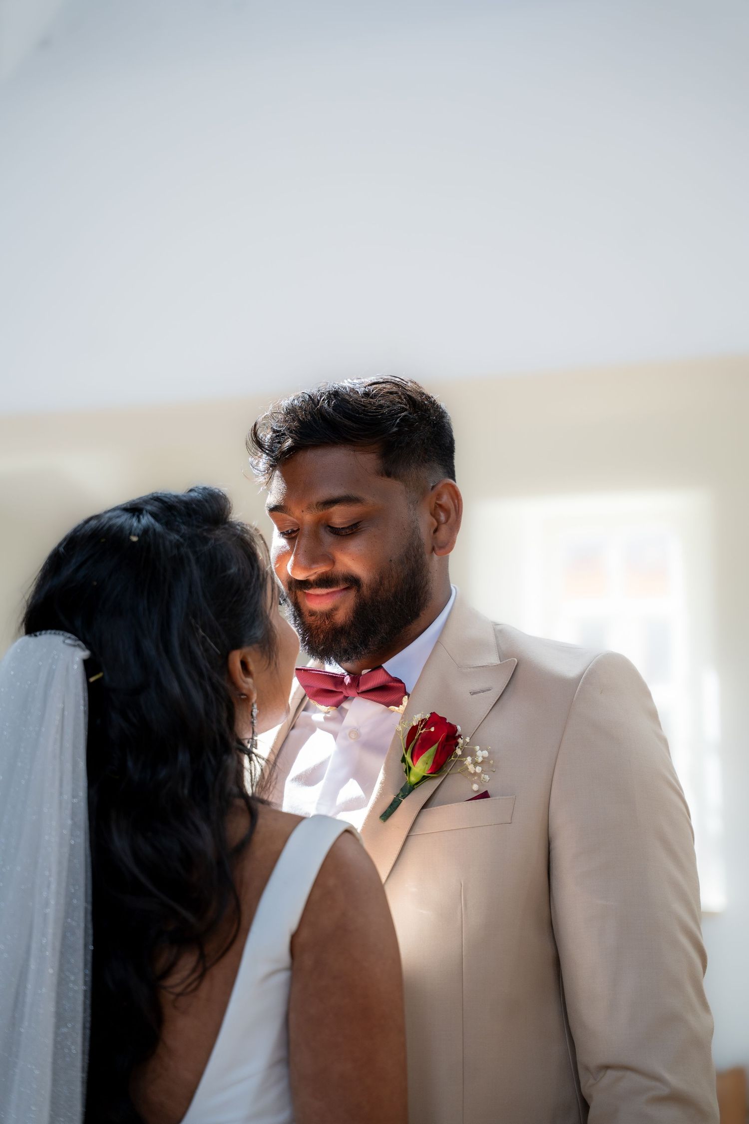 A groom in a beige suit with red bow tie and rose boutonniere smiles tenderly at his bride in a white wedding dress.