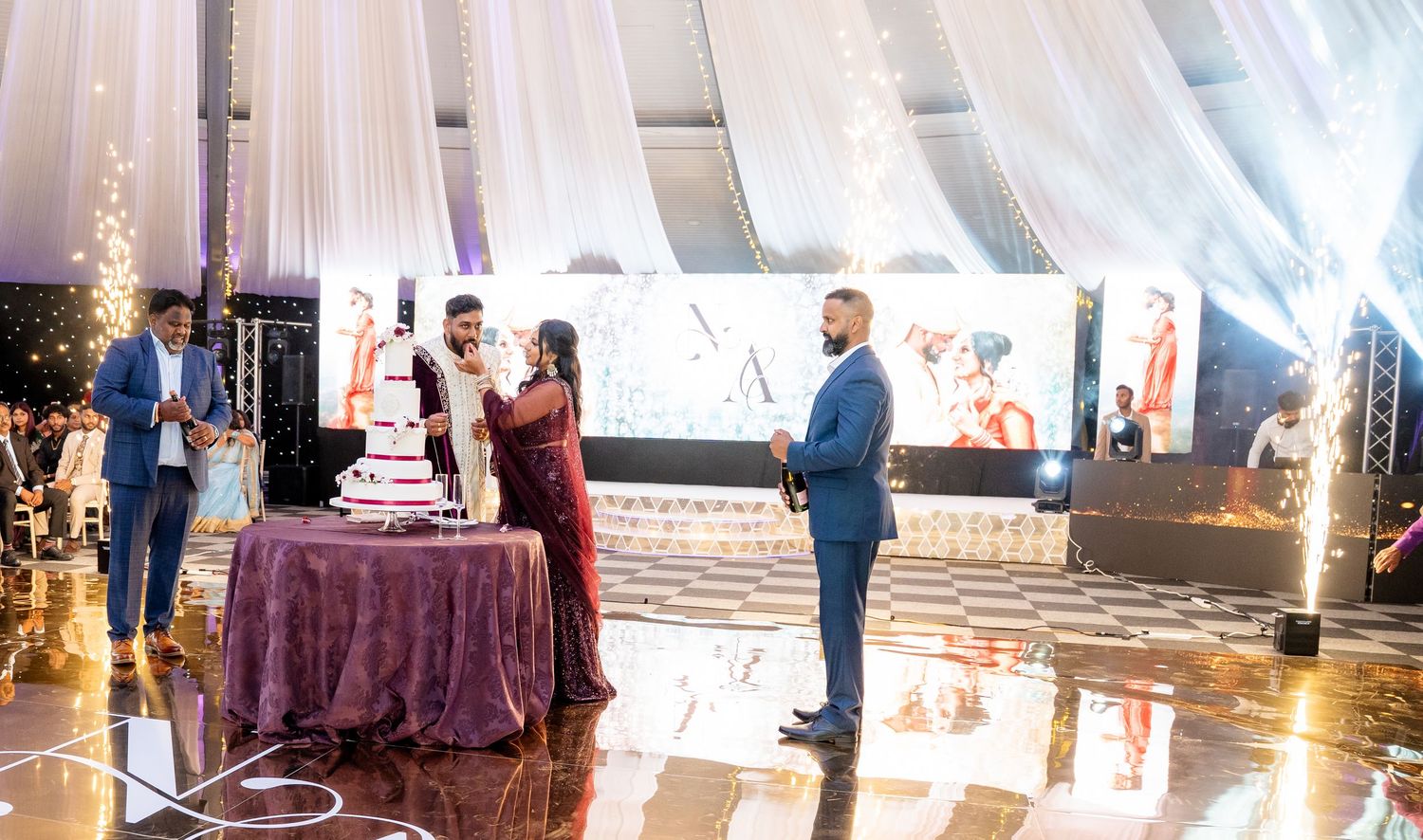 Indian couple cutting wedding cake surrounded by guests amid sparkling fireworks at glamorous reception.