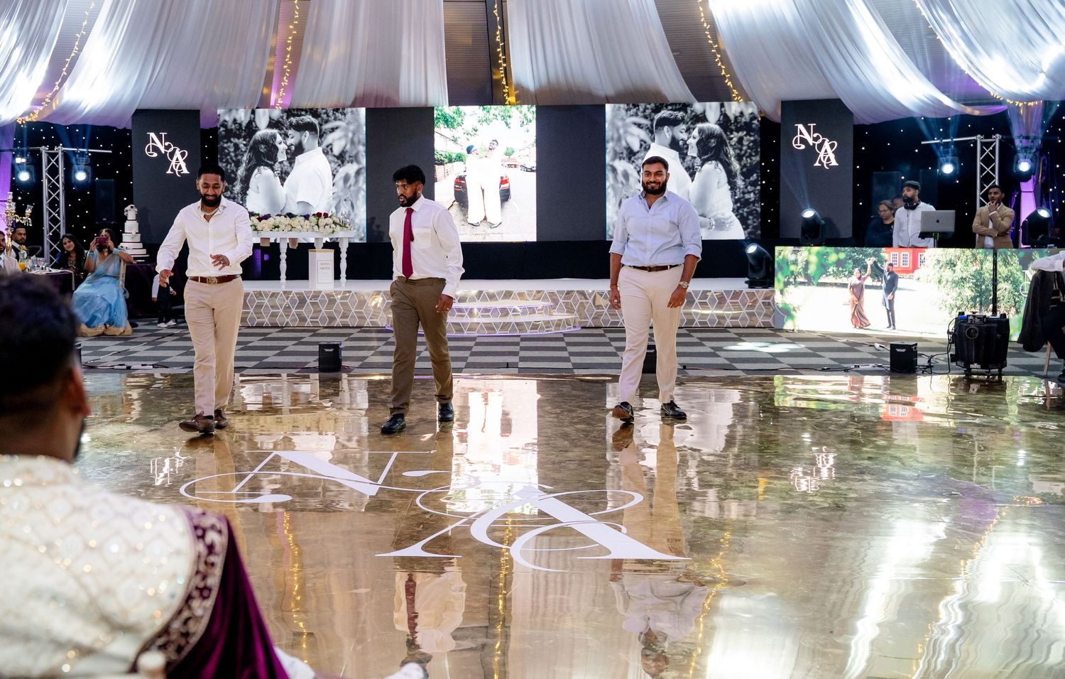 Three groomsmen in formal attire standing on glossy dance floor at an elegant Indian wedding venue.