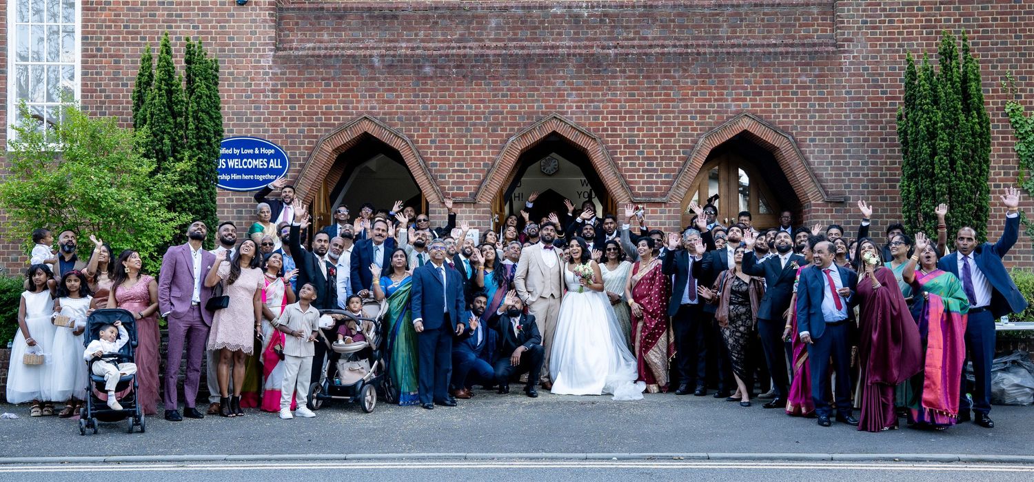Large wedding group photo outside a brick church, bride and groom surrounded by guests in formal attire on a sunny day.