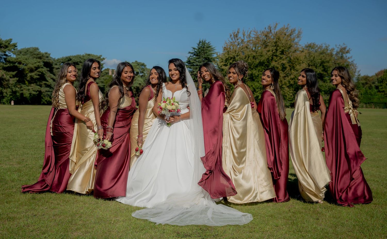 Bride in white gown poses with bridesmaids in burgundy and gold satin dresses on a sunny green lawn.