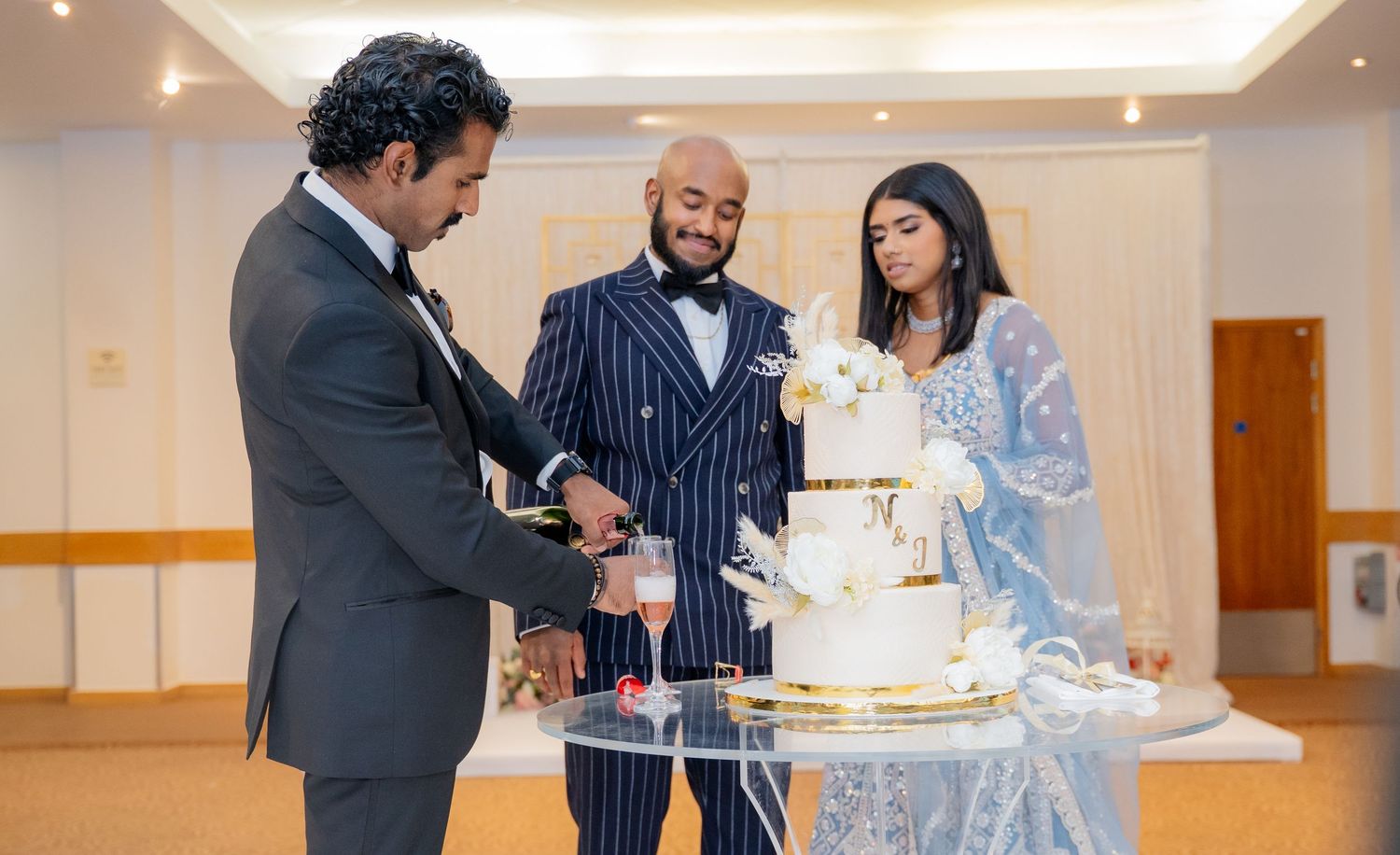 Three people cutting a white and gold tiered wedding cake at a formal reception.