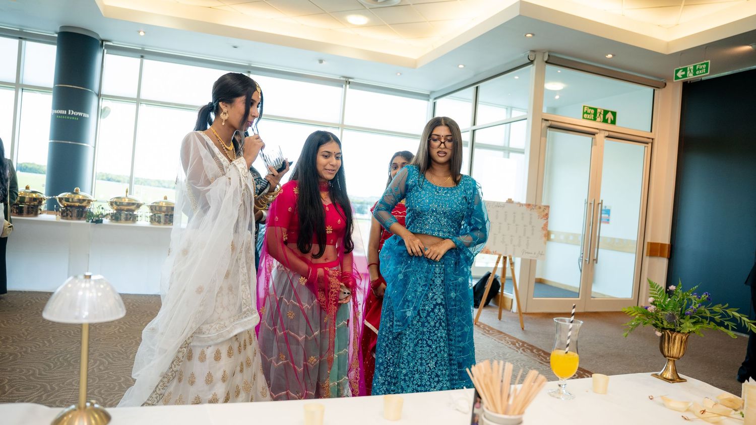 Three women wearing vibrant Indian traditional outfits standing together at a wedding event.