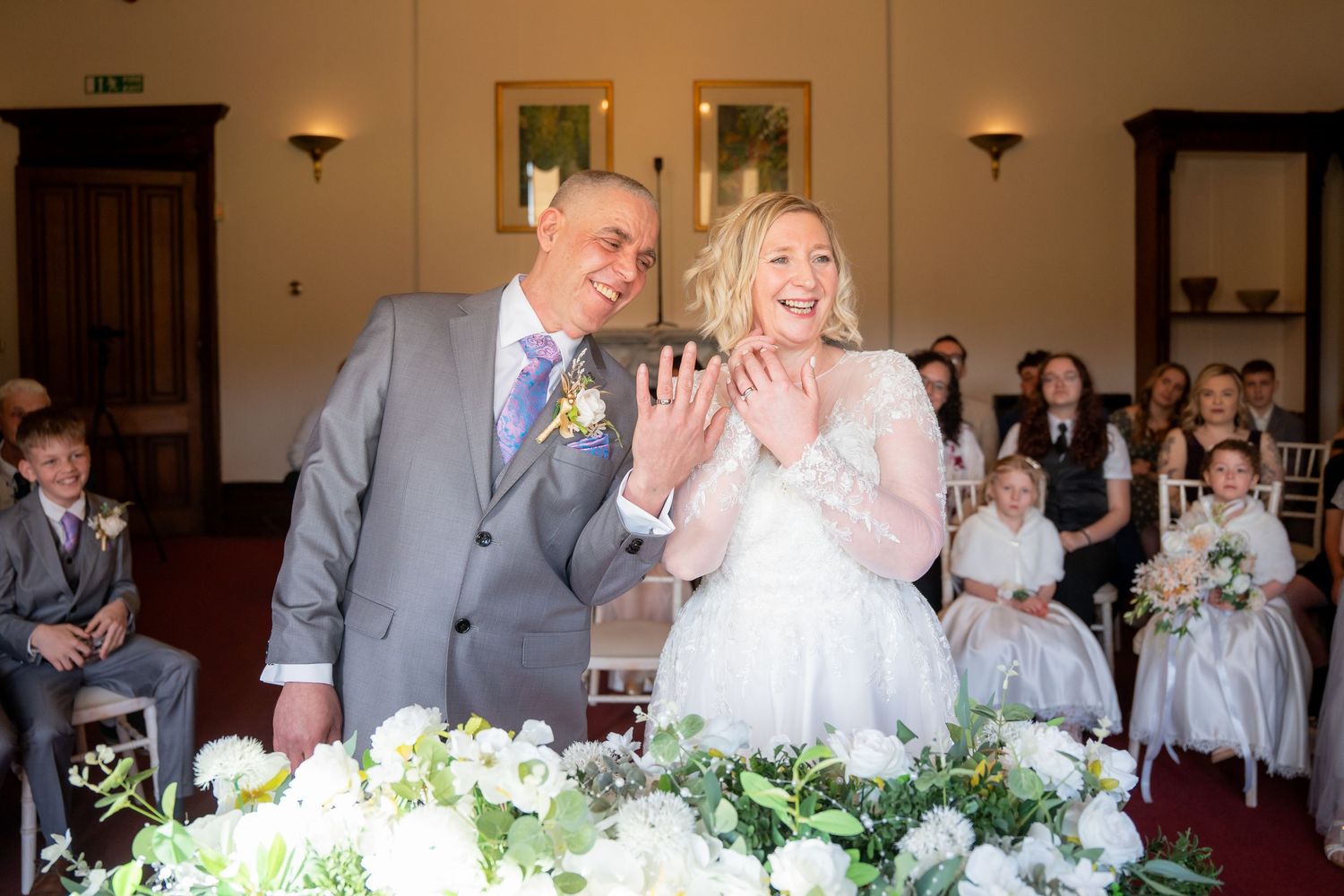 Joyful couple laughing at wedding ceremony altar surrounded by white floral arrangements and guests.