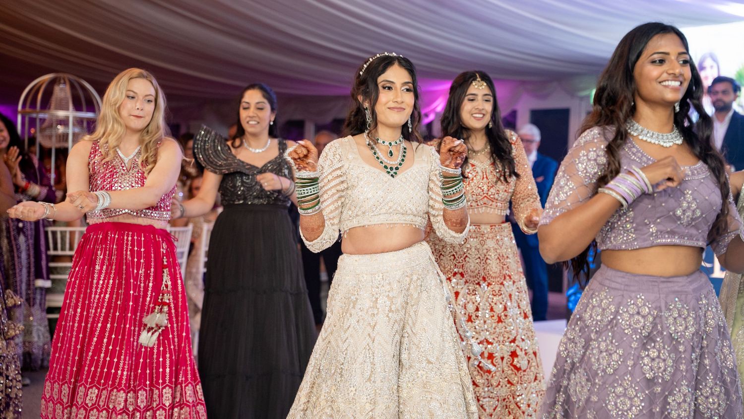 Bride in white lehenga dances with friends in colorful outfits at Indian wedding reception.