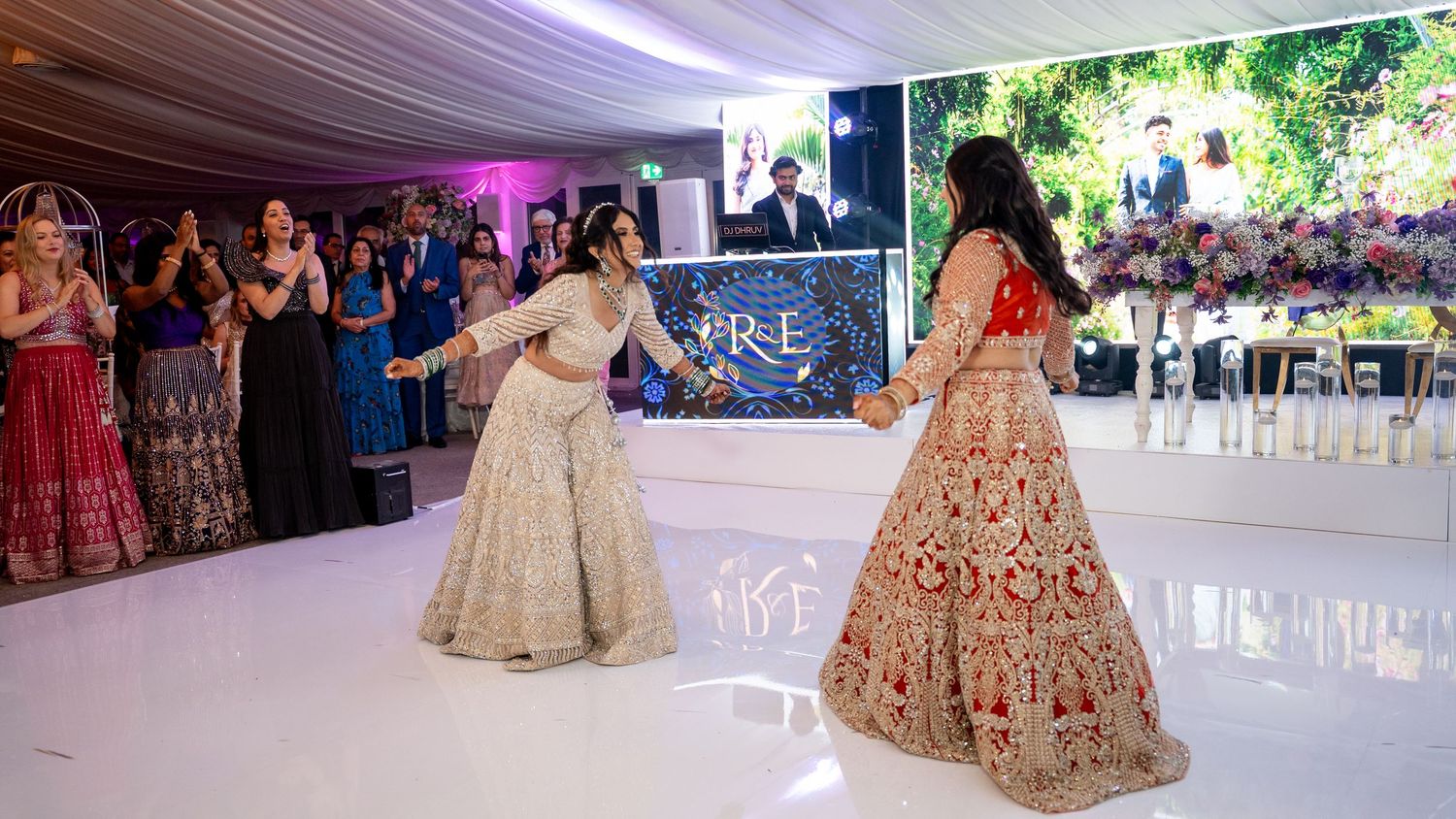 Women in elegant embroidered lehengas dancing at an indoor wedding reception with guests and decor in the background.
