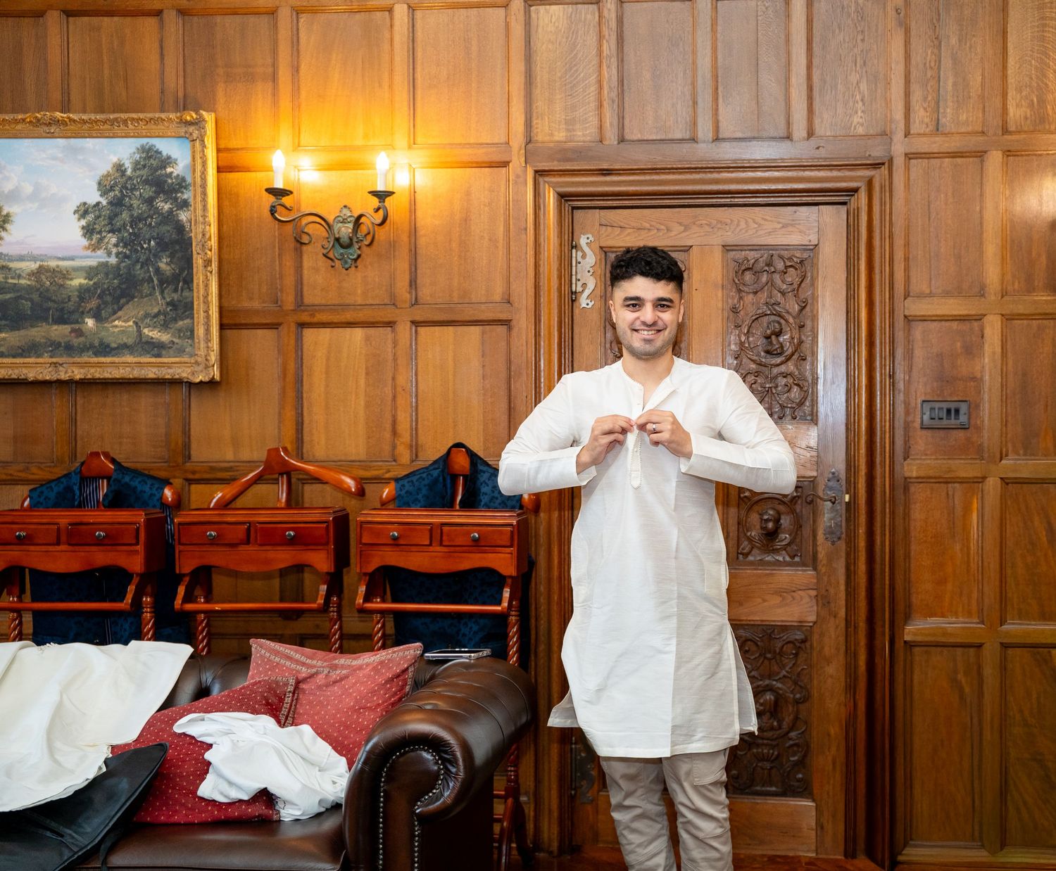 Young man in white kurta standing in elegant wood-paneled room with leather furniture and landscape painting.
