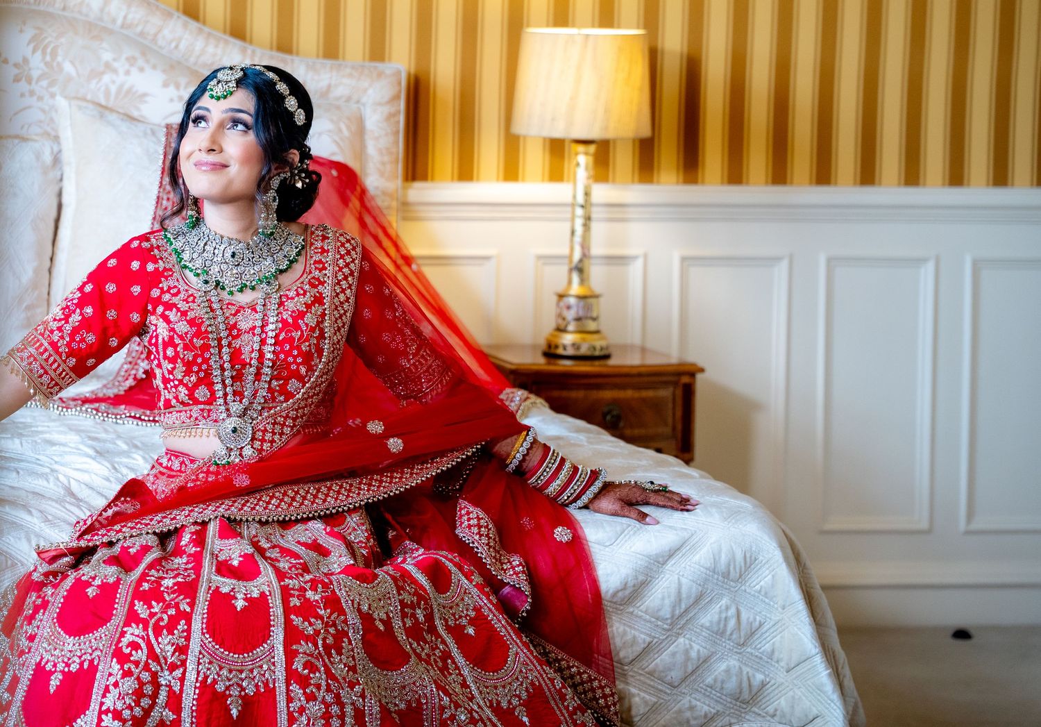 Indian bride in elaborate red embroidered lehenga gazes upward while seated on white bed.
