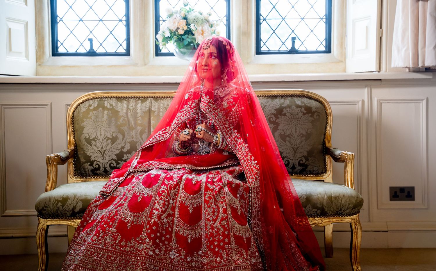 Indian bride in red lehenga seated on gold sofa holding bouquet near window.