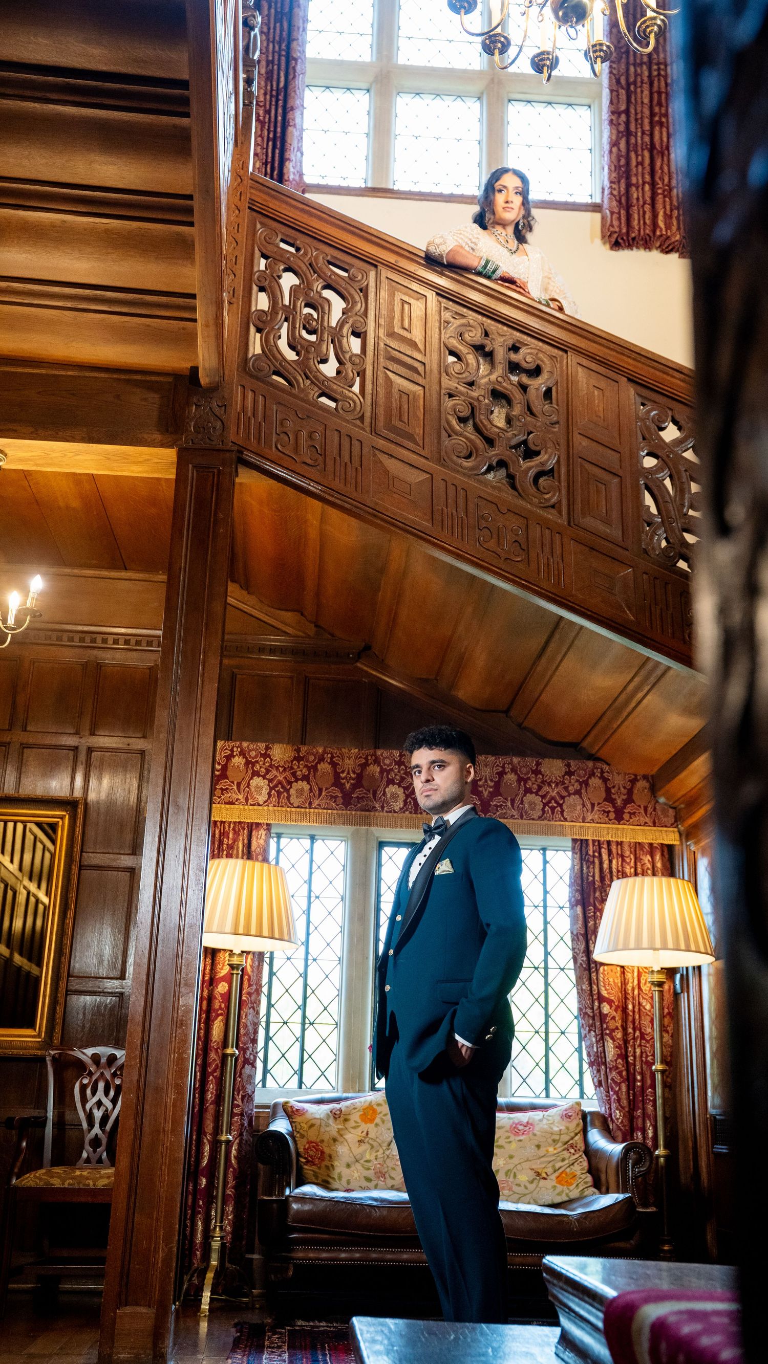 Groom in teal suit stands in ornate wood-paneled hall as bride watches from carved balcony above.