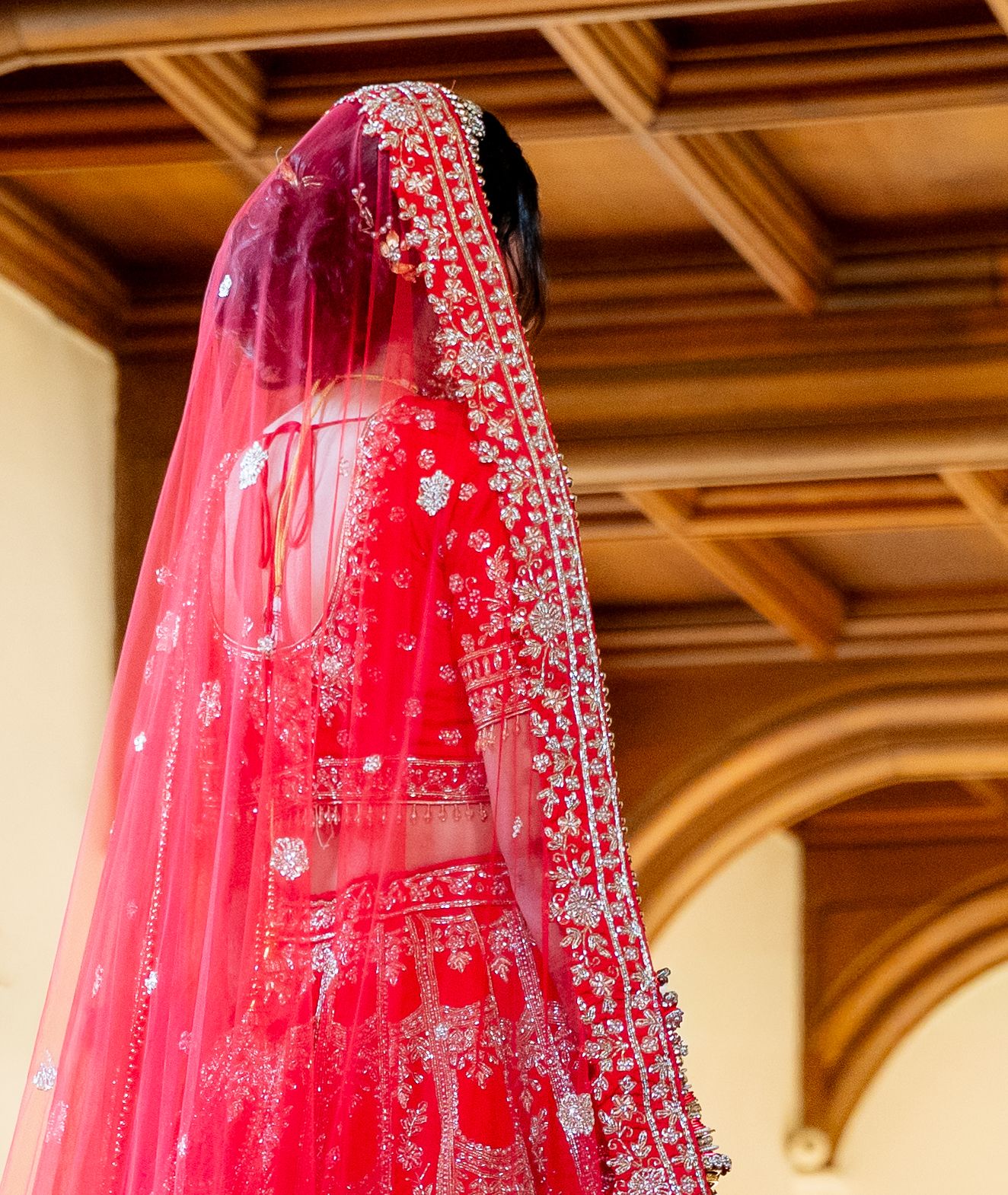Stunning red bridal lehenga with intricate silver embroidery and dupatta veil, photographed from behind in elegant venue.